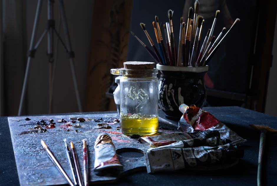 A cluttered artist's table filled with paint tubes, brushes, and a glass jar.