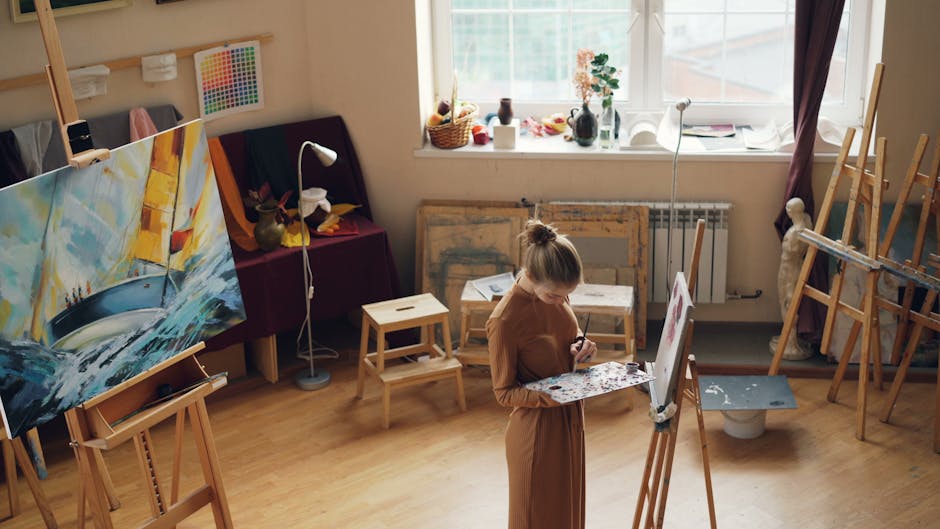 A female artist painting in a brightly lit studio surrounded by art supplies and paintings.