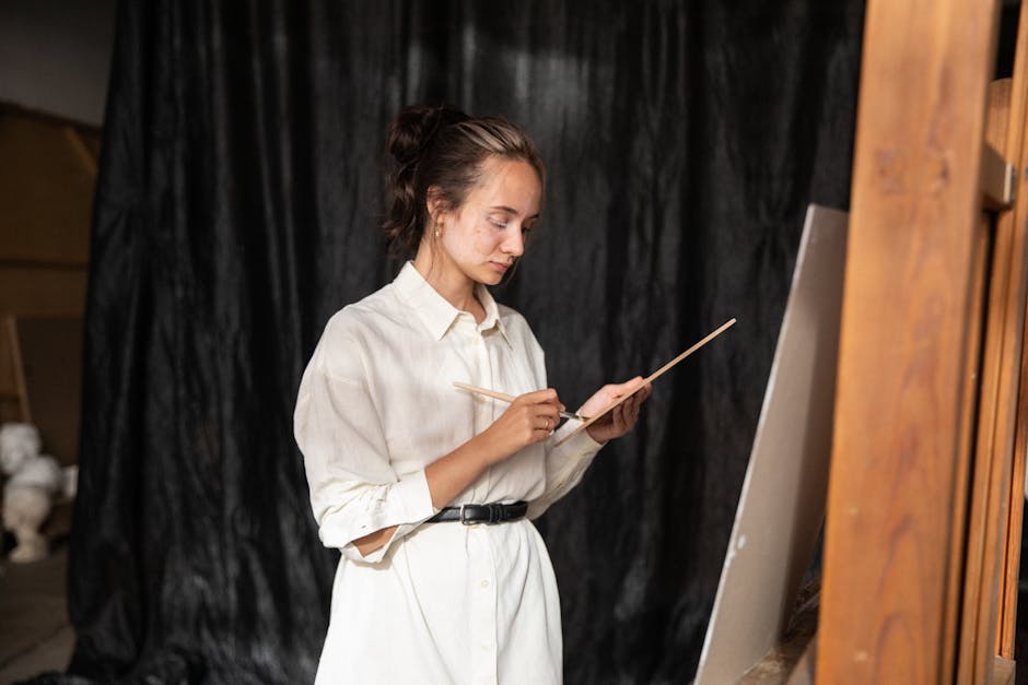 A woman artist in a studio wearing a button-up shirt, focused on painting with a palette and brush.