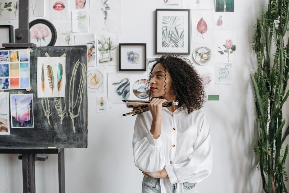 Female artist with curly hair holding paintbrushes in a studio with artwork on the walls.