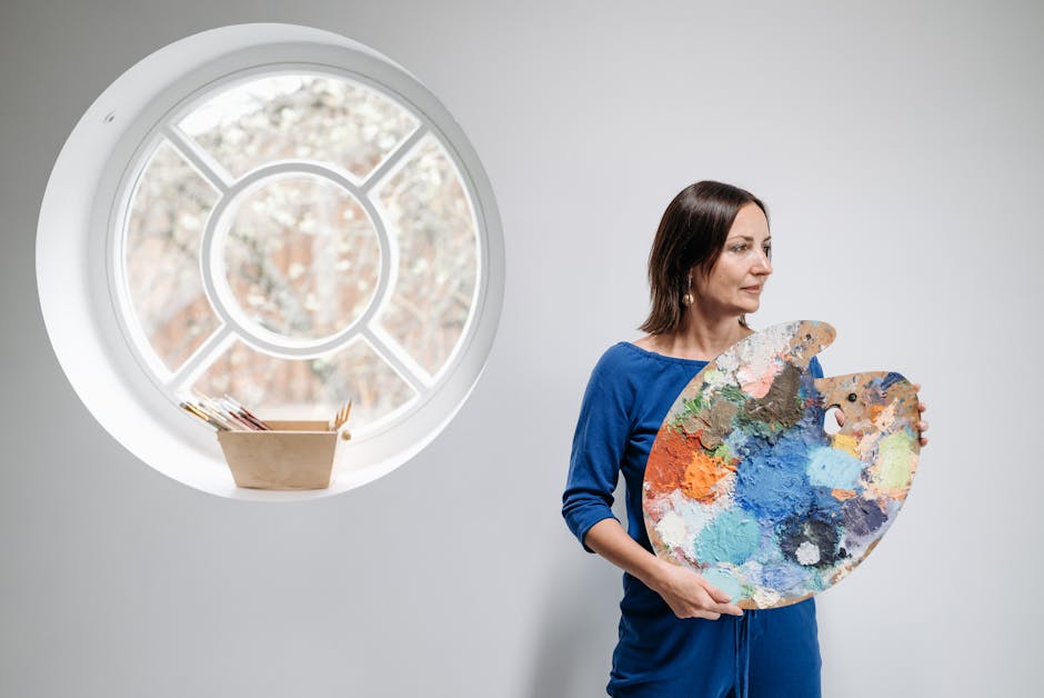 Woman in blue dress holding vibrant artist palette by a round window indoors.
